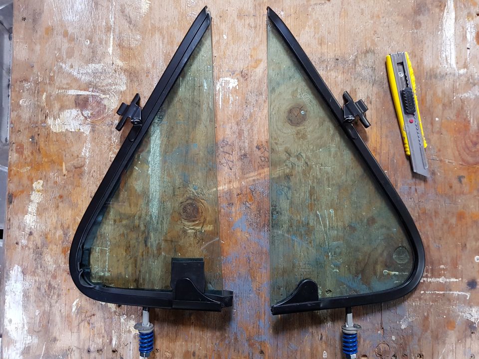 two triangular vent windows with black trim and mounting hardware, next to a utility knife on a wooden surface