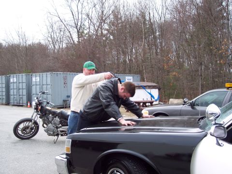 2001 Ford Crown Victoria - A staged traffic-stop scene in a gravel lot: one man leans over the hood of a black sedan while another in a green cap stands behind him. A…