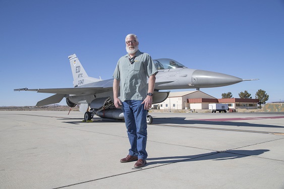 Fighter jet on tarmac with a man standing in front. The jet is gray with markings on the tail