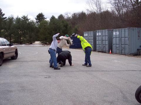 2001 Ford Crown Victoria - Four men act out a scuffle on an asphalt lot lined with gray shipping containers and a dumpster, one wearing a yellow high-visibility jacke…