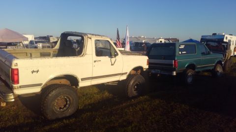 1988 Ford Bronco - Two Ford Broncos, one white with a removed roof and the other green, are parked at an outdoor event.