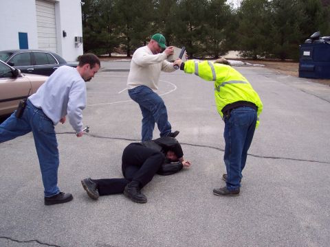 2001 Ford Crown Victoria - Three men stand over a fourth lying on the pavement in what looks like a staged arrest or takedown drill, one wearing a yellow high-visibil…