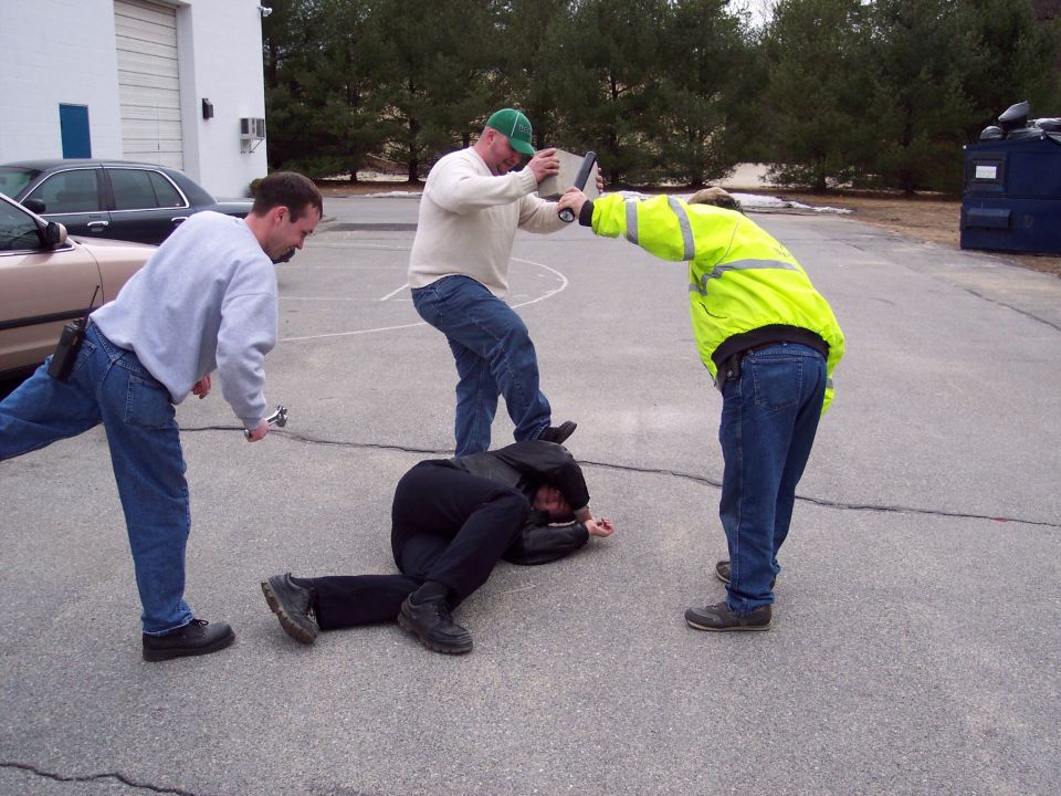 Staged arrest drill on pavement with three men over a man on the ground, tan sedan parked at left