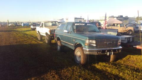 1988 Ford Bronco - Two trucks, a green Bronco and a white pickup, are parked in a grassy field with RVs in the background.