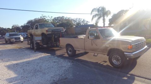 1988 Ford Bronco - A tan 1987 Ford F-250 tows a lifted truck on a trailer, with a GMC SUV visible in the background.