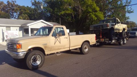 1988 Ford Bronco - A tan Ford F-Series truck tows a lifted truck on a trailer down a residential street.
