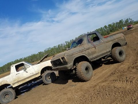 1988 Ford Bronco - Two heavily muddied trucks, including a 1978 Ford Bronco, navigate a muddy off-road course under a bright blue sky.