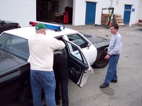 2001 Ford Crown Victoria - Two men lean into the open rear door of a black-and-white police cruiser parked in a shop lot while a third man in a gray sweatshirt watche…