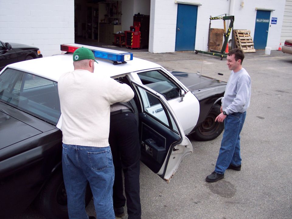 Men working at open rear door of black-and-white police cruiser with roof lightbar in shop lot