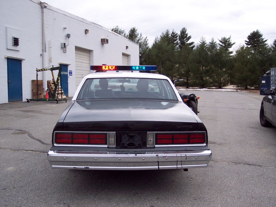 Black and white police car with light bar, rear view. Scratched chrome bumper