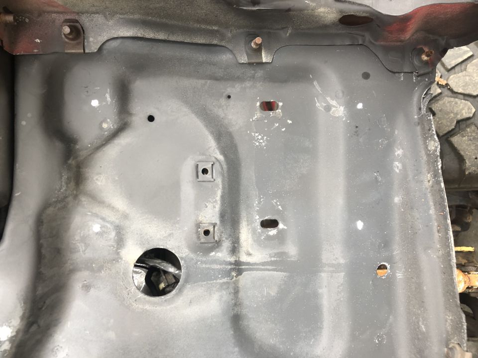 Undercarriage of a vehicle, showing gray painted metal with some rust and white paint specks. A tire is visible on the right