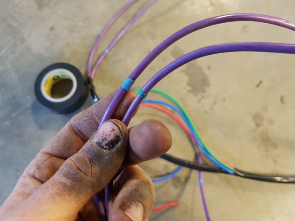 Hand holding purple electrical wires with blue and green tape markers. Roll of black electrical tape in background