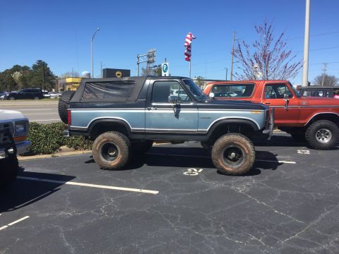 1979 Ford Bronco - A lifted blue Ford Bronco is parked next to another Bronco in a parking lot on a sunny day.