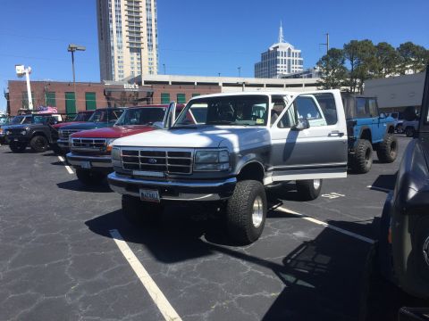 1979 Ford Bronco - A line of lifted trucks and SUVs are parked in a lot, with a white and grey Ford Bronco in the foreground.