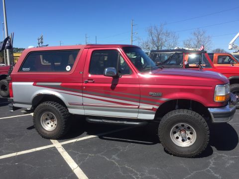1979 Ford Bronco - A red and silver Ford Bronco is parked in a lot with other vehicles under a clear blue sky.