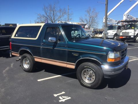 1979 Ford Bronco - A green Ford Bronco with a tan stripe and a white removable top is parked in a lot with utility trucks in the background.
