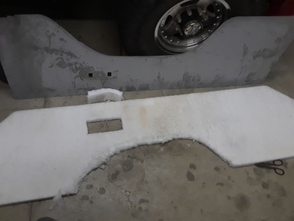 Gray vehicle interior panel with cutouts and white insulation material, next to a silver wheel