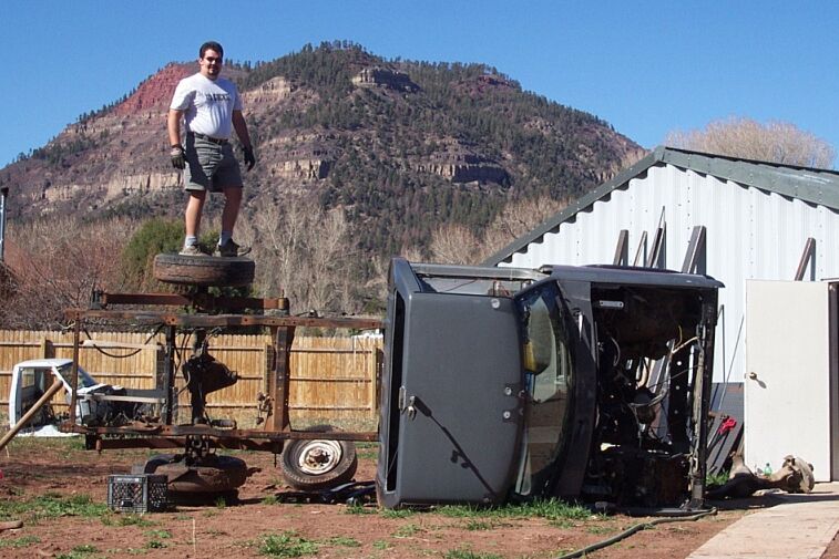 Stripped truck cab tipped on its side in a dirt yard with a man standing on the bare chassis