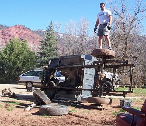 1990 Ford Bronco - A man stands triumphantly on the tire of an overturned vehicle, with another car parked nearby.