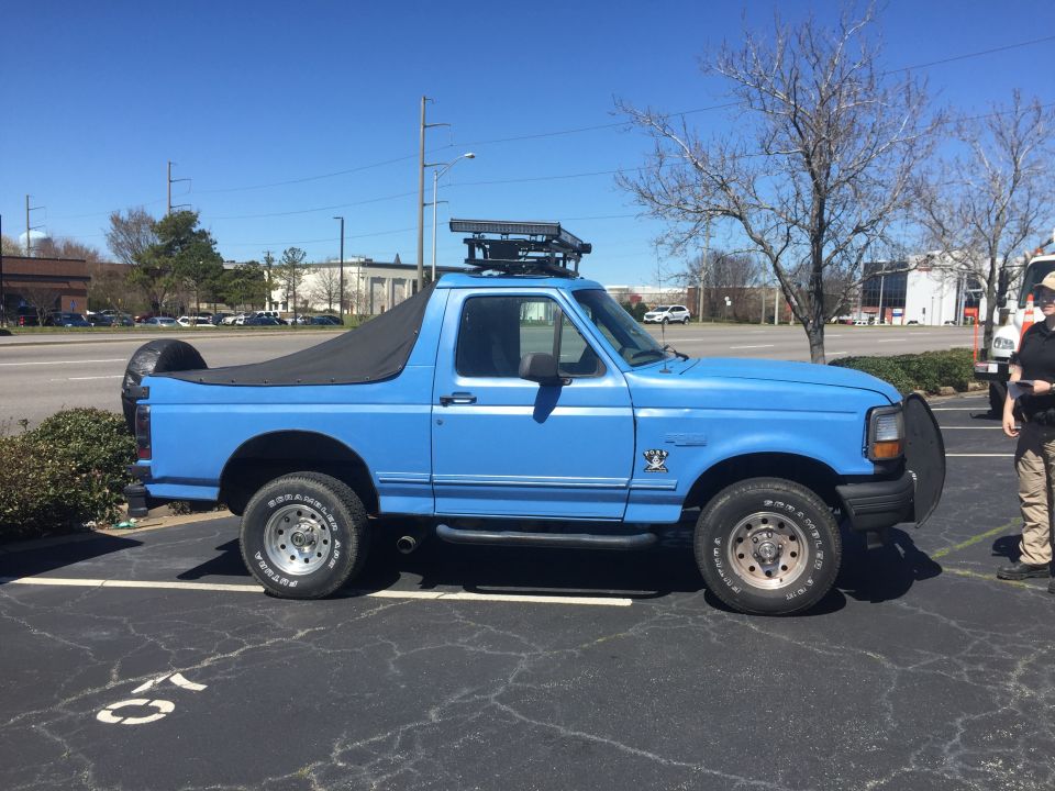 Blue Ford Bronco with a black soft top, roof rack, and spare tire on the back. The vehicle has a bull bar on the front