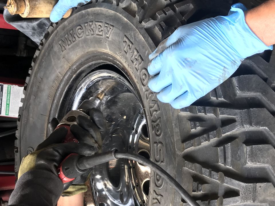 Hands in blue gloves and a work glove using a tool on a Mickey Thompson tire valve stem