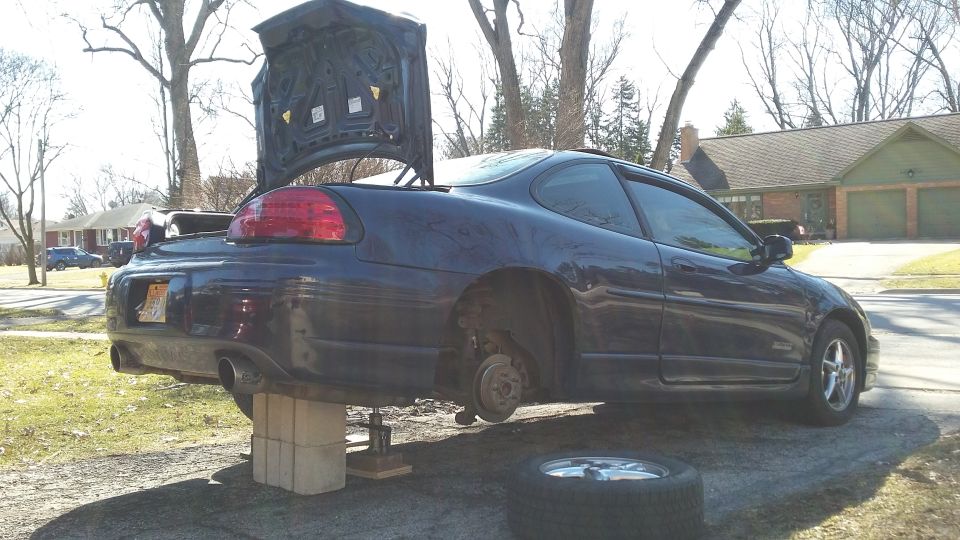 Dark blue 2000 Pontiac Grand Prix coupe with trunk open and rear lifted on blocks, front wheel missing
