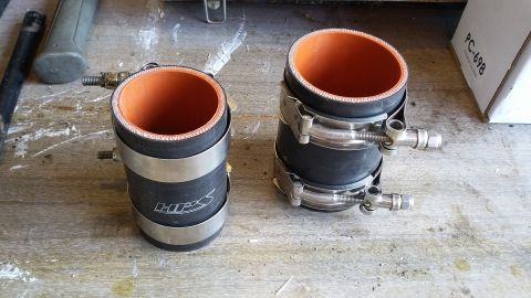 Two black silicone hoses with orange interiors and metal clamps on a wooden surface. One hose has "HPS" logo