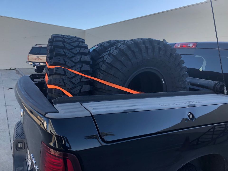 Four large off-road tires secured with orange straps in the bed of a black pickup truck. Another pickup truck is visible in the background
