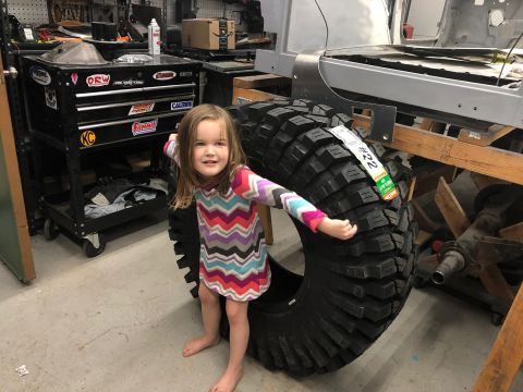 1992 Jeep Wrangler - A young girl poses next to a massive tire in a garage setting, with tools and vehicle parts visible.