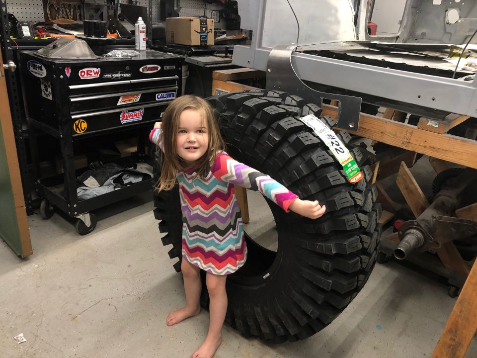Girl in a colorful dress stands by a large off-road tire in a workshop. A toolbox is to the left