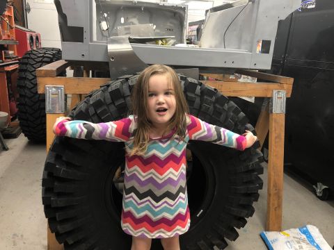 1992 Jeep Wrangler - A young girl stands in front of a large tire and the unfinished body of a vehicle in a workshop.