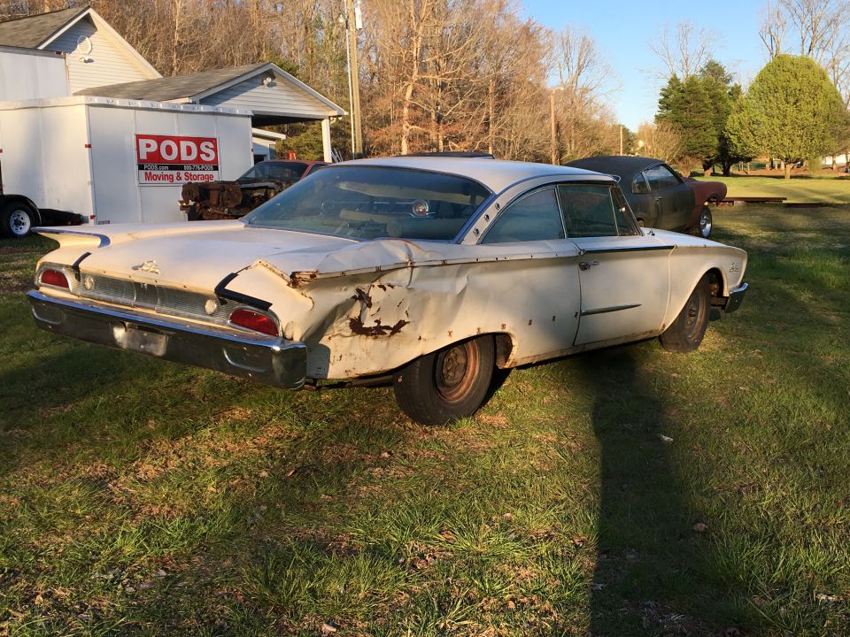 Old white Ford coupe with significant rear damage, parked on grass. Another car is visible behind it