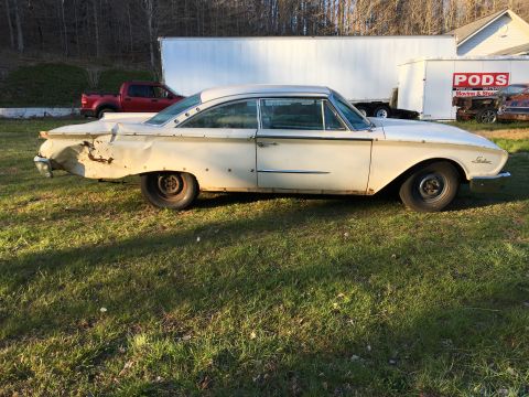 1960 Ford Starliner - A white 1960 Ford Starliner coupe sits in a grassy field, showing extensive damage to its rear passenger side.