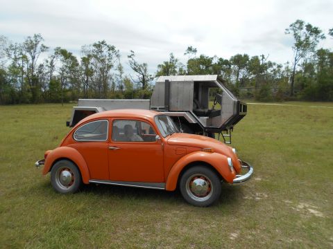 1992 Ford Bronco - An orange Volkswagen Beetle sits on a grassy field beside a unique metallic trailer structure.