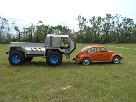 1992 Ford Bronco - A custom truck tows a classic orange Volkswagen Beetle across a grassy field under a cloudy sky.
