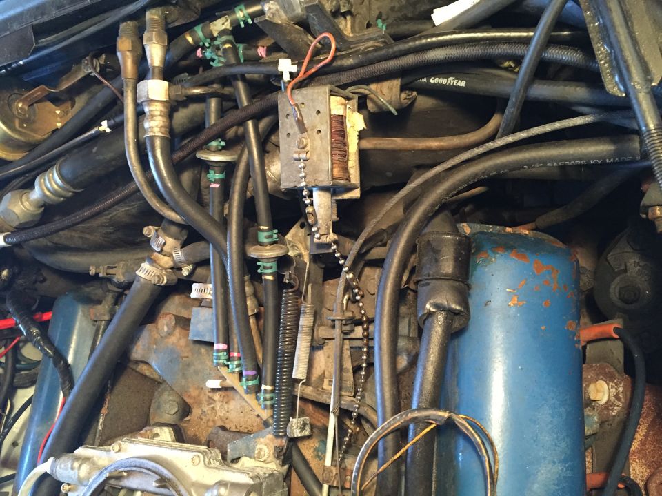 Close-up of a car's engine bay, showing fuel lines, a carburetor, and various hoses and wires