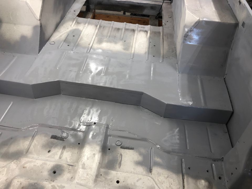 Gray painted vehicle floor with visible ribs and mounting holes, showing the transmission tunnel and seat mounts