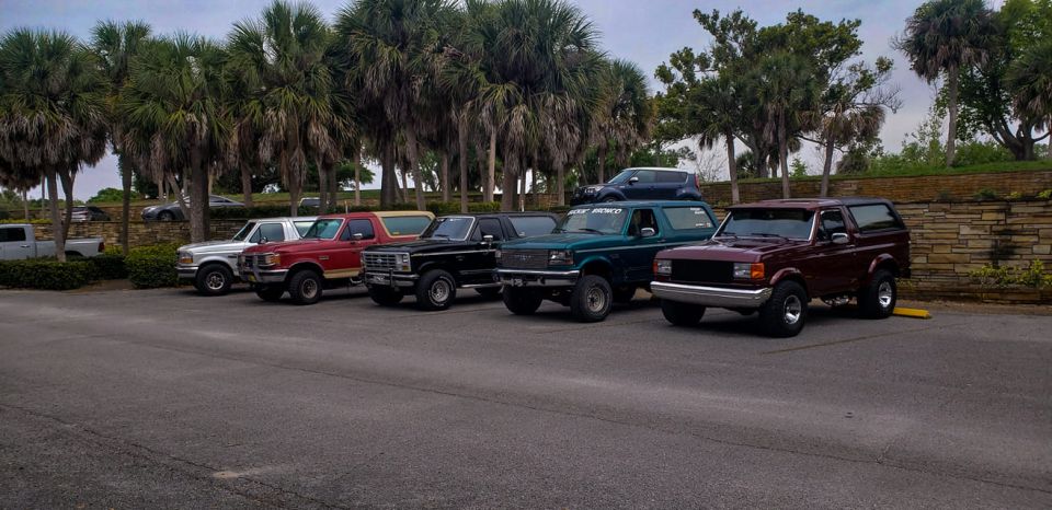 Several Ford Broncos and trucks parked in a lot, including a maroon Bronco from the early 90s