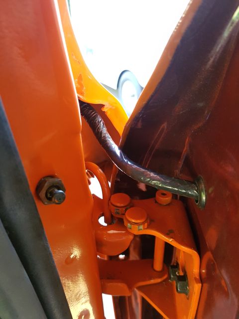 1978 Ford Bronco - Detail of the door hinge mechanism and wiring on an orange vehicle, with a glimpse of another vehicle in the background.