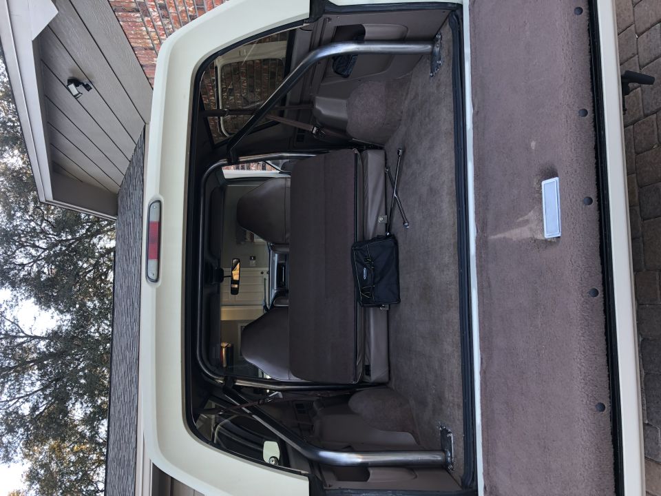 White 1995 Ford Bronco interior with roll cage, folded rear seat, and jack. Rear cargo area is carpeted