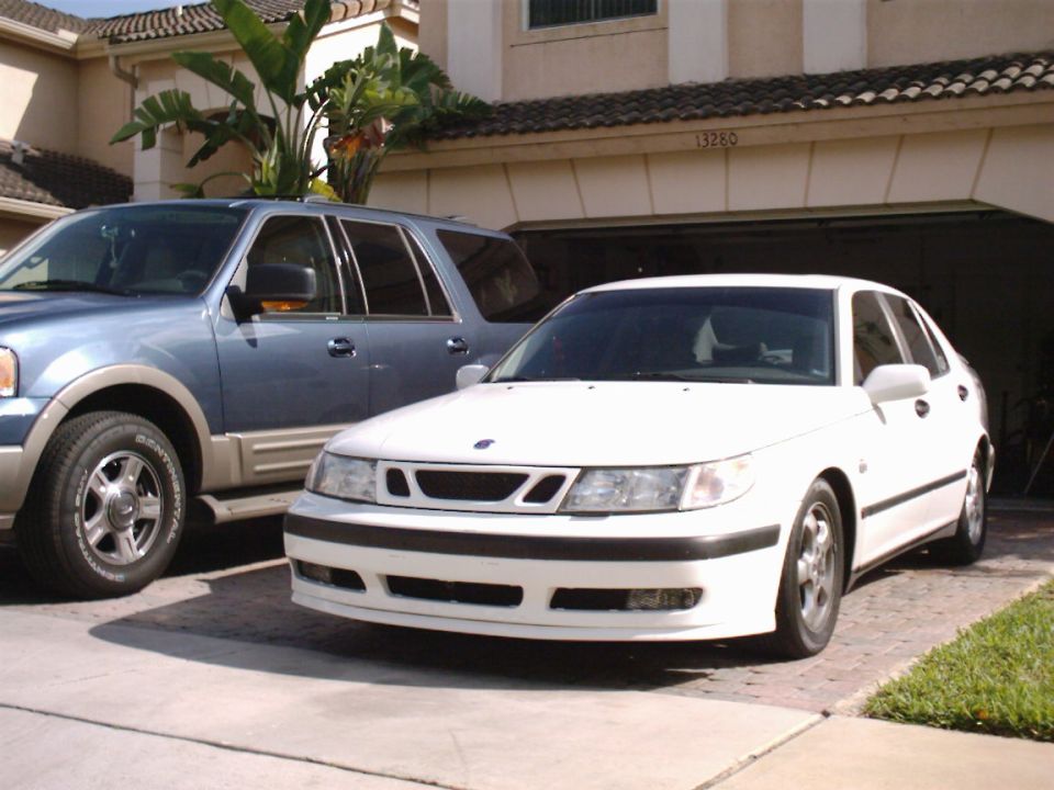 White Saab 9-5 sedan parked in driveway next to blue SUV, front 3/4 view
