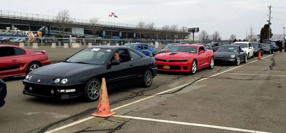 Black coupe with racing numbers on windshield in line with other cars at a track