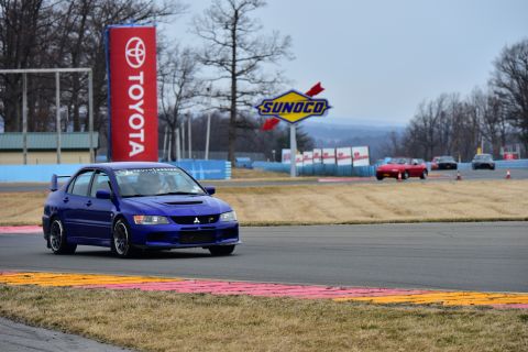 2006 Mitsubishi Evolution - A blue 2006 Mitsubishi Lancer Evolution races on a track, with other vehicles visible in the background and sponsor signs overhead.