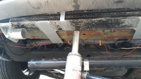 Car's underside with a jack lifting it near the radiator and exhaust pipe. Wires and rusty frame are visible