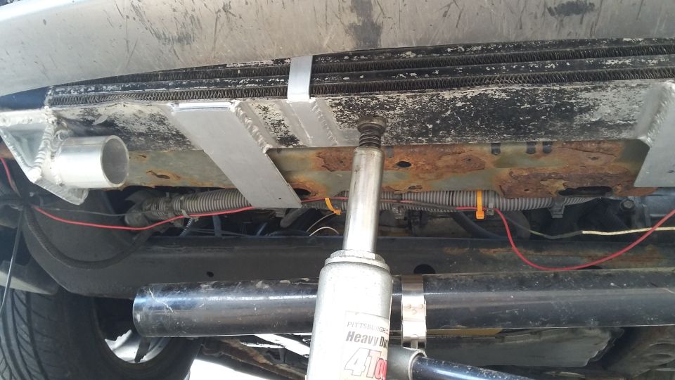 Car's underside with a jack lifting it near the radiator and exhaust pipe. Wires and rusty frame are visible