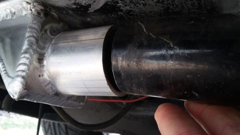 Close-up of a metal exhaust pipe connection with visible welds and a black pipe section. A finger is near the