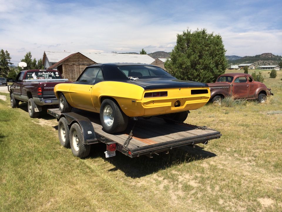 Yellow 1969 Chevrolet Camaro on a trailer being towed by a maroon Dodge pickup truck. Another old car is parked in the background