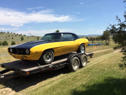 1969 Chevrolet Camaro - A yellow and black coupe is transported on a trailer through a grassy, rural landscape under a partly cloudy sky.