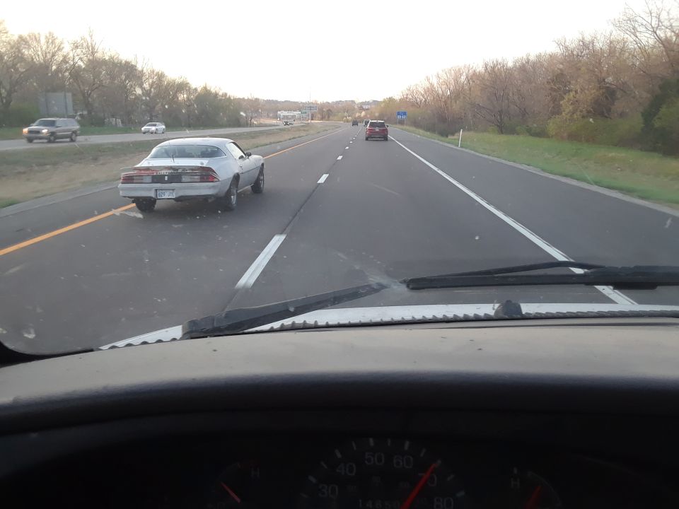 White 1980s Chevrolet Camaro driving on highway, rear view