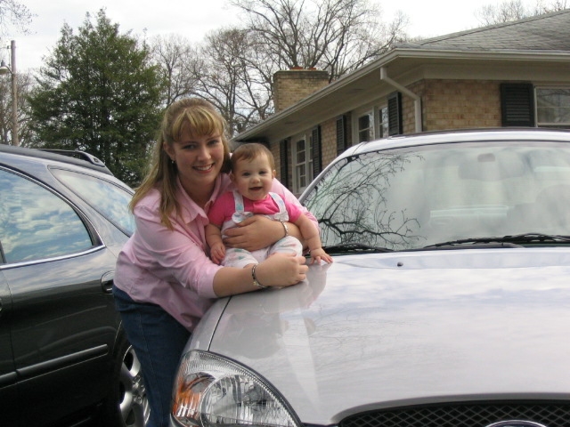 Silver 2004 Ford Taurus front end with a woman holding a baby on the hood
